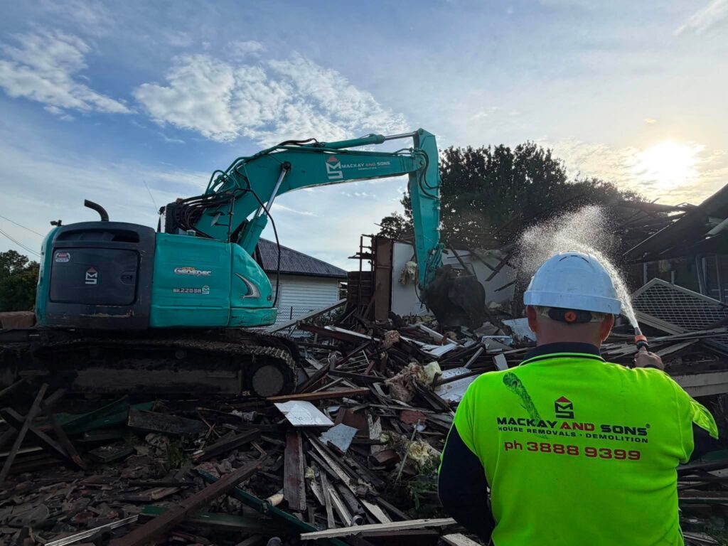 Excavator demolishing a house with a worker in hi-vis and a water spray controlling dust